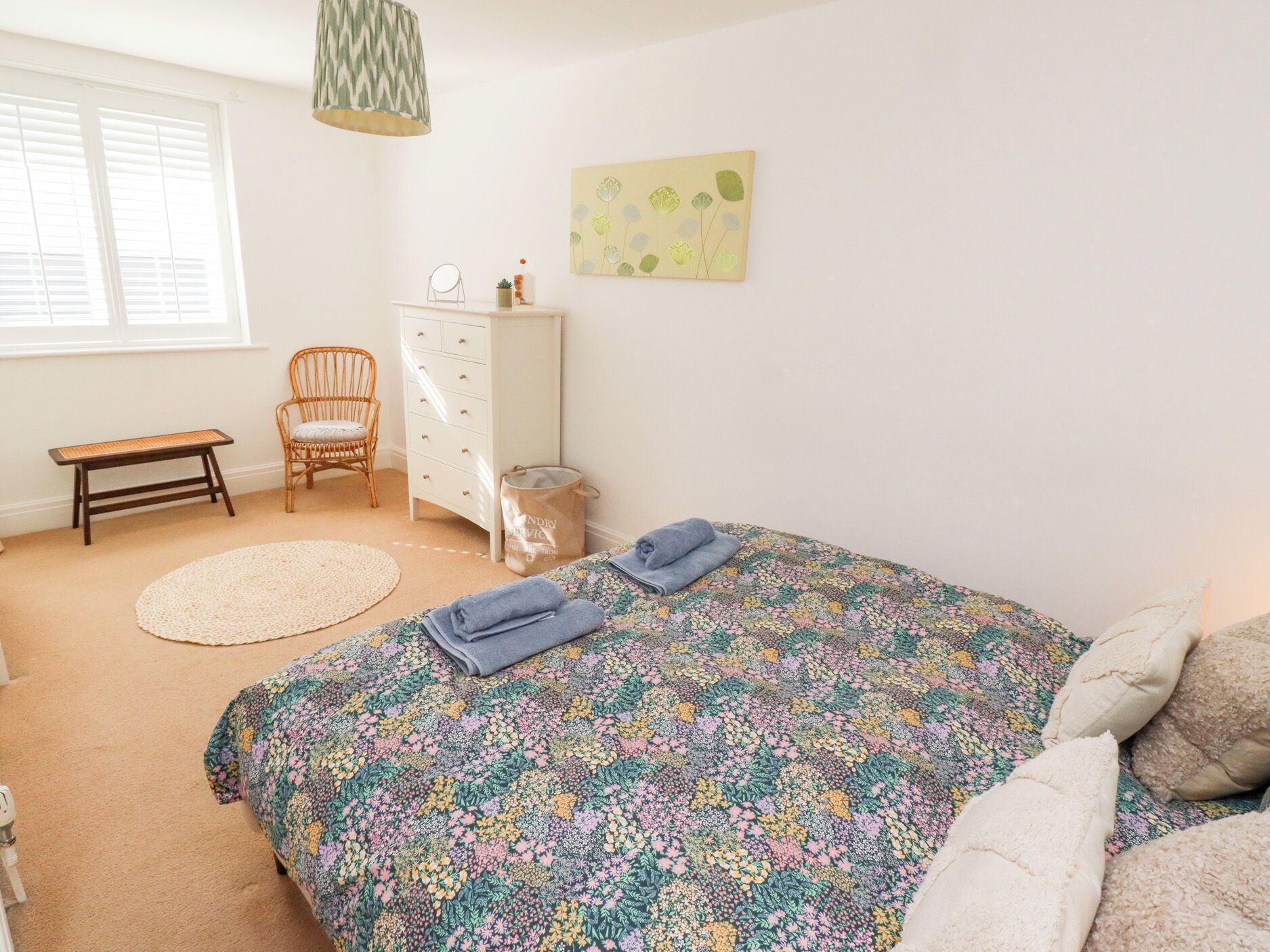A bedroom with a floral bedspread, a white dresser, a wicker chair, a bench, and a window with blinds.