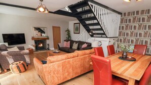 A modern living room with a sofa, a dining table with chairs, a fireplace, a staircase, and a bookshelf.