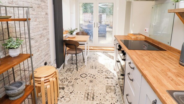 A compact kitchen with a wooden countertop, a stove, and a sink. There is a dining area with a table and chairs visible through a glass door.