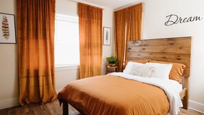 A bedroom with a wooden headboard, a large bed with a brown bedspread, and two windows with orange curtains. - Beautiful One Bedroom Apartment in The Neighbor of the Art (Rochester)