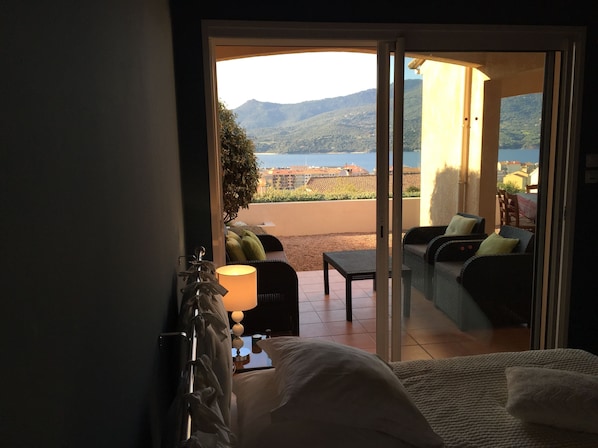 Une chambre comprenant un lit, une table de chevet, une lampe et donnant sur un balcon aménagé avec du mobilier d’extérieur, offrant une vue sur un paysage pittoresque.