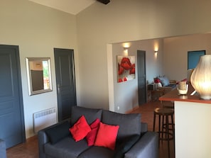 Un salon avec un canapé gris et des coussins rouges, un coin repas avec une table et des chaises, et une cuisine à l’arrière-plan.
