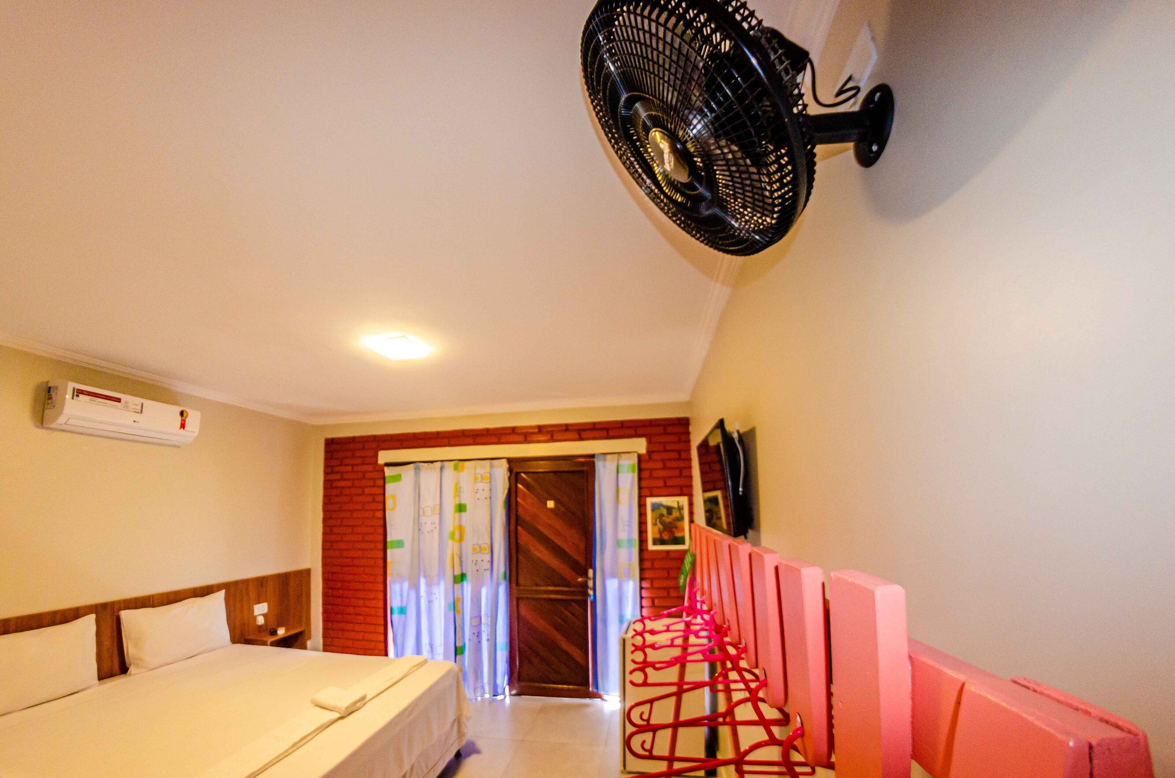 A hotel room with a bed, a wall-mounted fan, a door with curtains, and a TV on the wall.