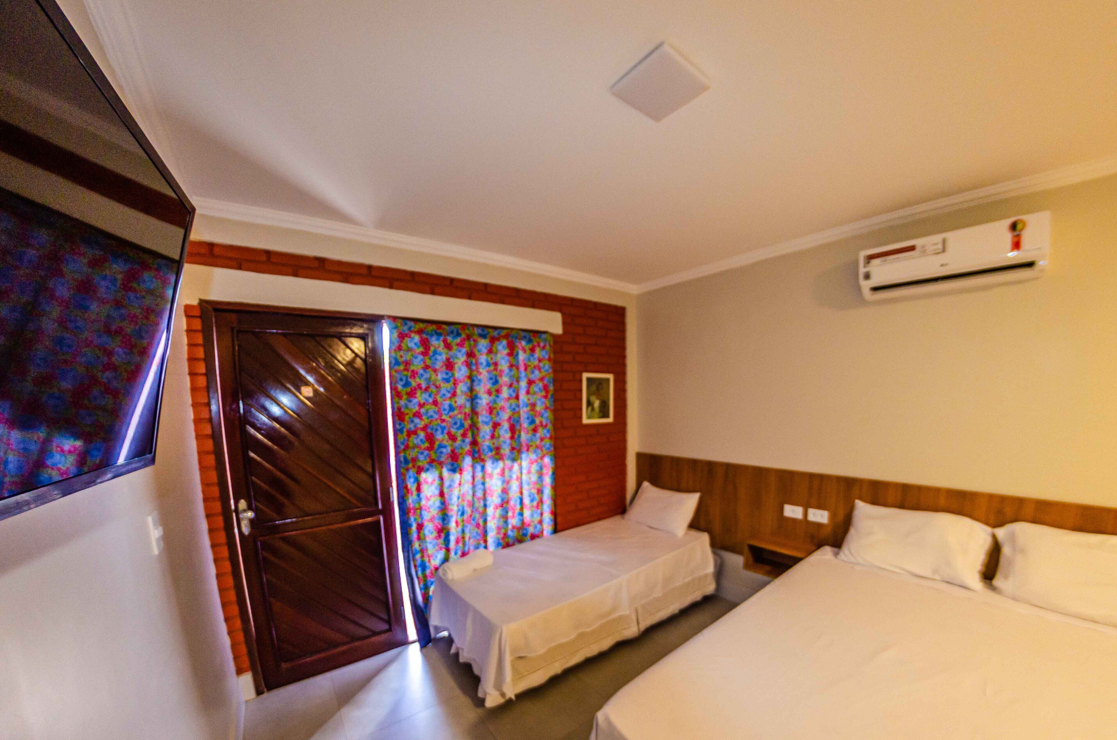A hotel room with two single beds, a wooden headboard, a large window with a floral curtain, and an air conditioning unit on the wall.