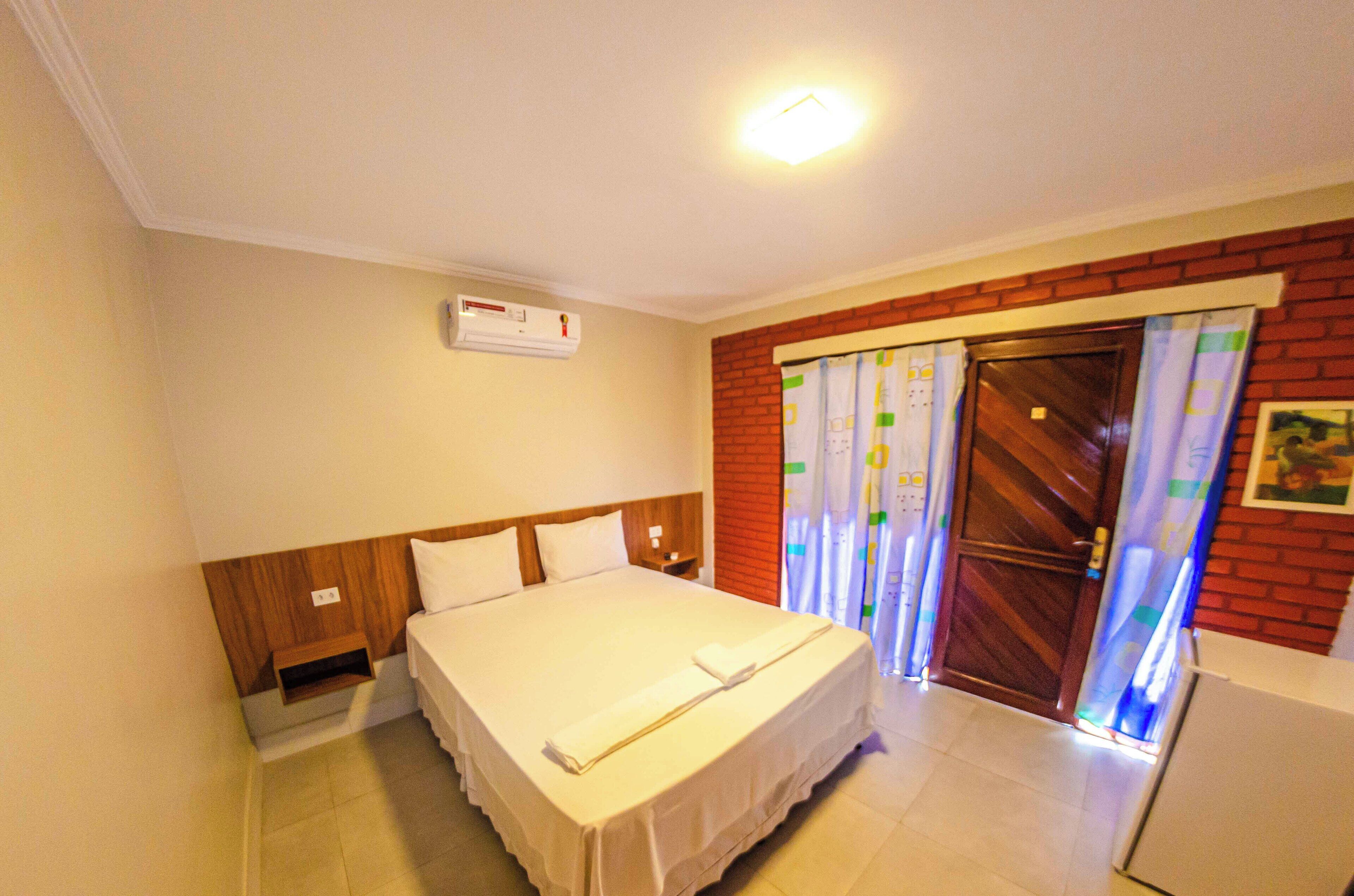 Habitación de hotel con una cama grande, cabecera de madera, mesita de noche, aire acondicionado de pared, puerta con cortina y una pequeña mesa.