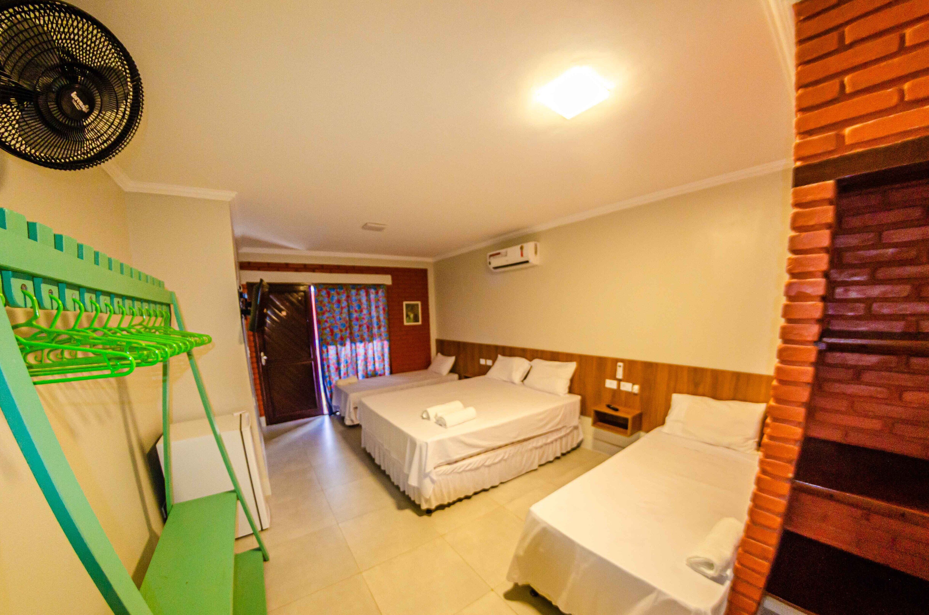 Habitación de hotel con dos camas, una silla plegable verde, un ventilador y una chimenea de ladrillo.