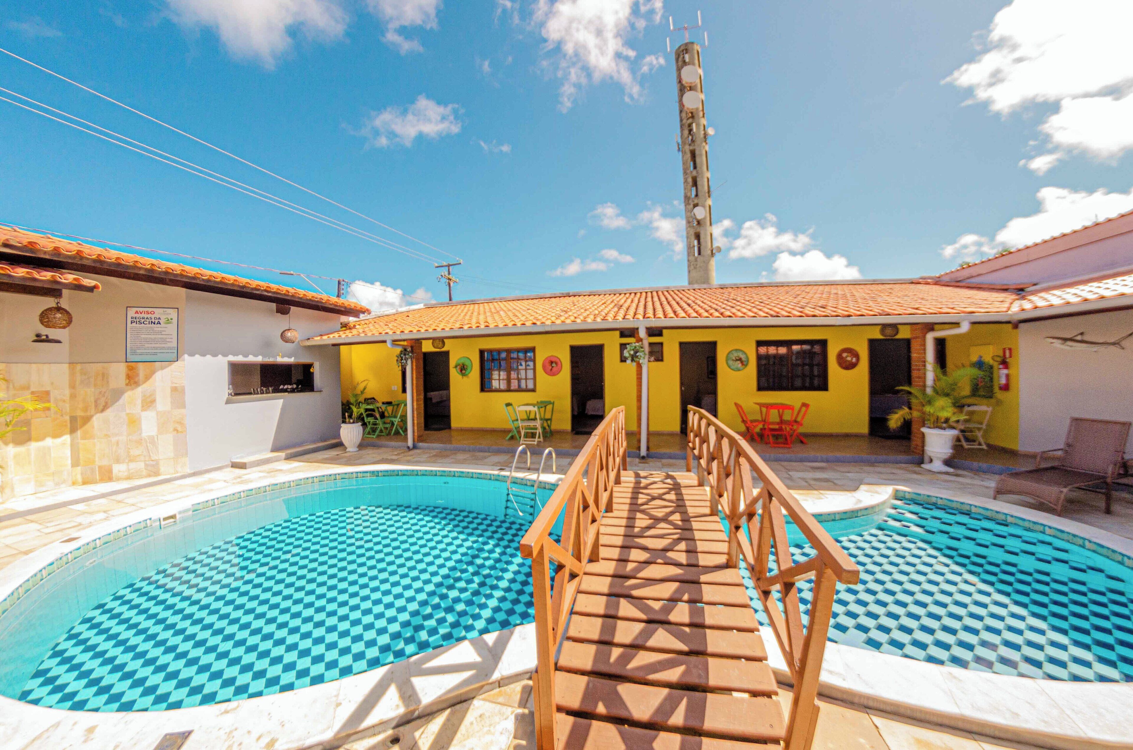 Un espace piscine comprenant deux piscines, une terrasse en bois et un bâtiment aux murs jaunes et au toit recouvert de tuiles.