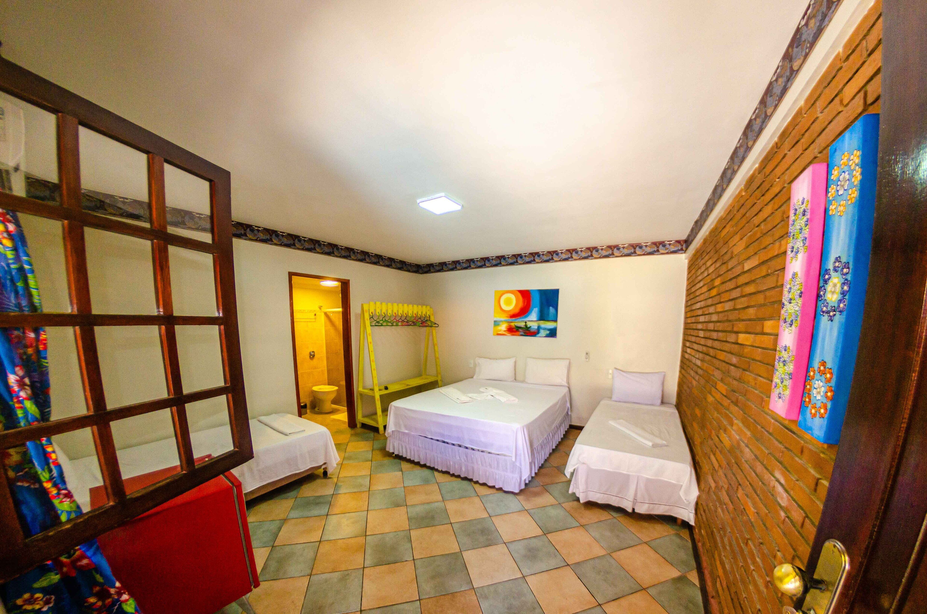 A hotel room with two single beds, a small table, a chair, a wooden door, and a colorful painting on the wall.