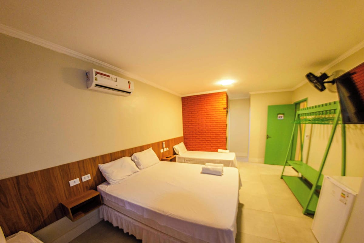 Habitación de hotel con cama, mesitas de noche, unidad de aire acondicionado, una puerta verde y un refrigerador pequeño.