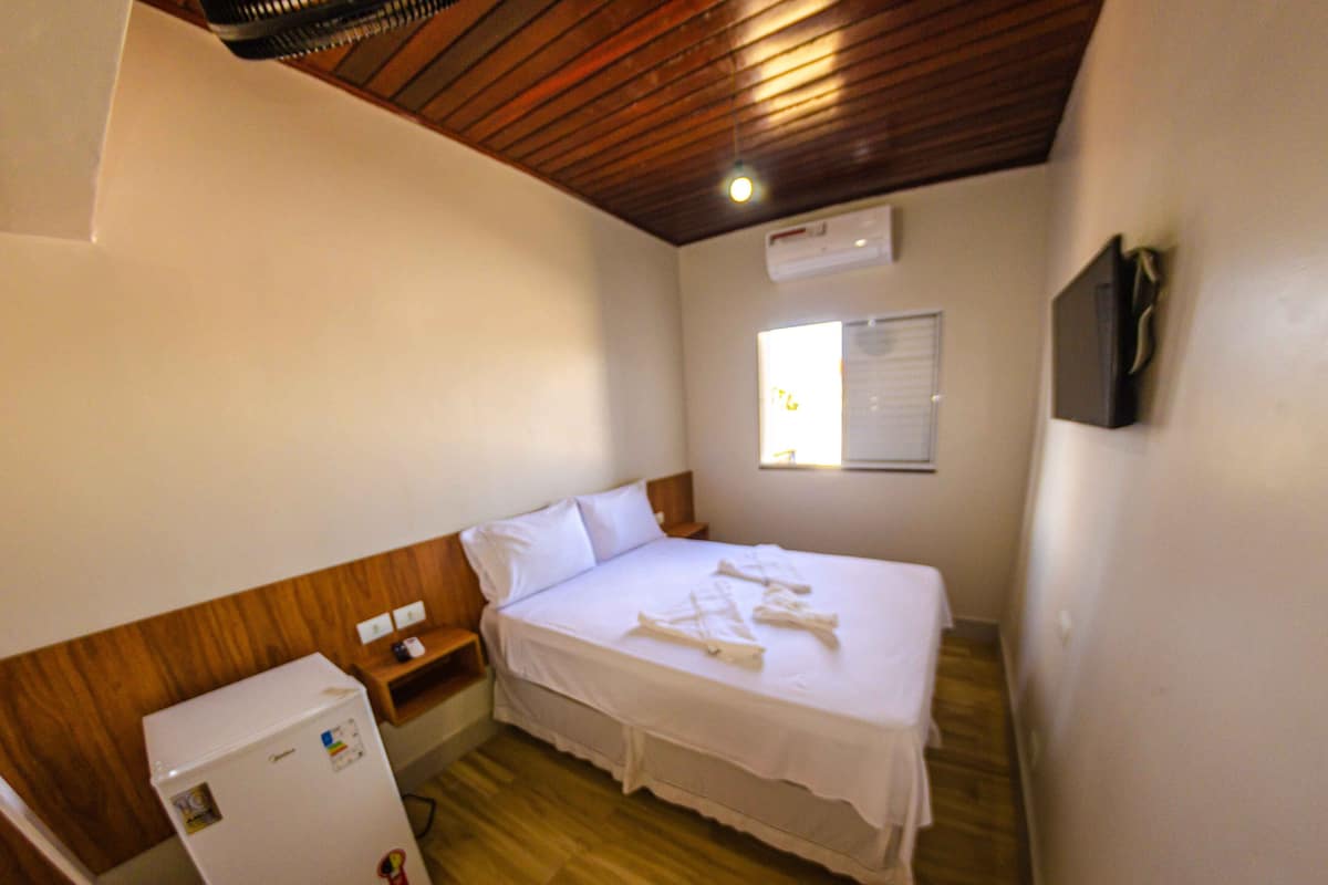 Un dormitorio con techo de madera, una cama con ropa de cama blanca, una mesita de noche pequeña, un televisor montado en la pared y una ventana con persianas.