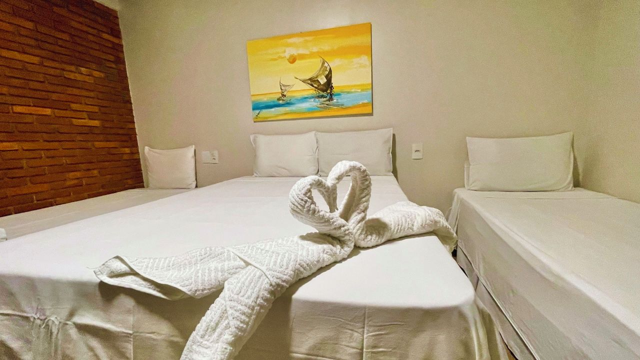 Habitación de hotel con una cama grande, un cuadro de un atardecer sobre el agua en la pared y una toalla doblada en forma de corazón sobre la cama.