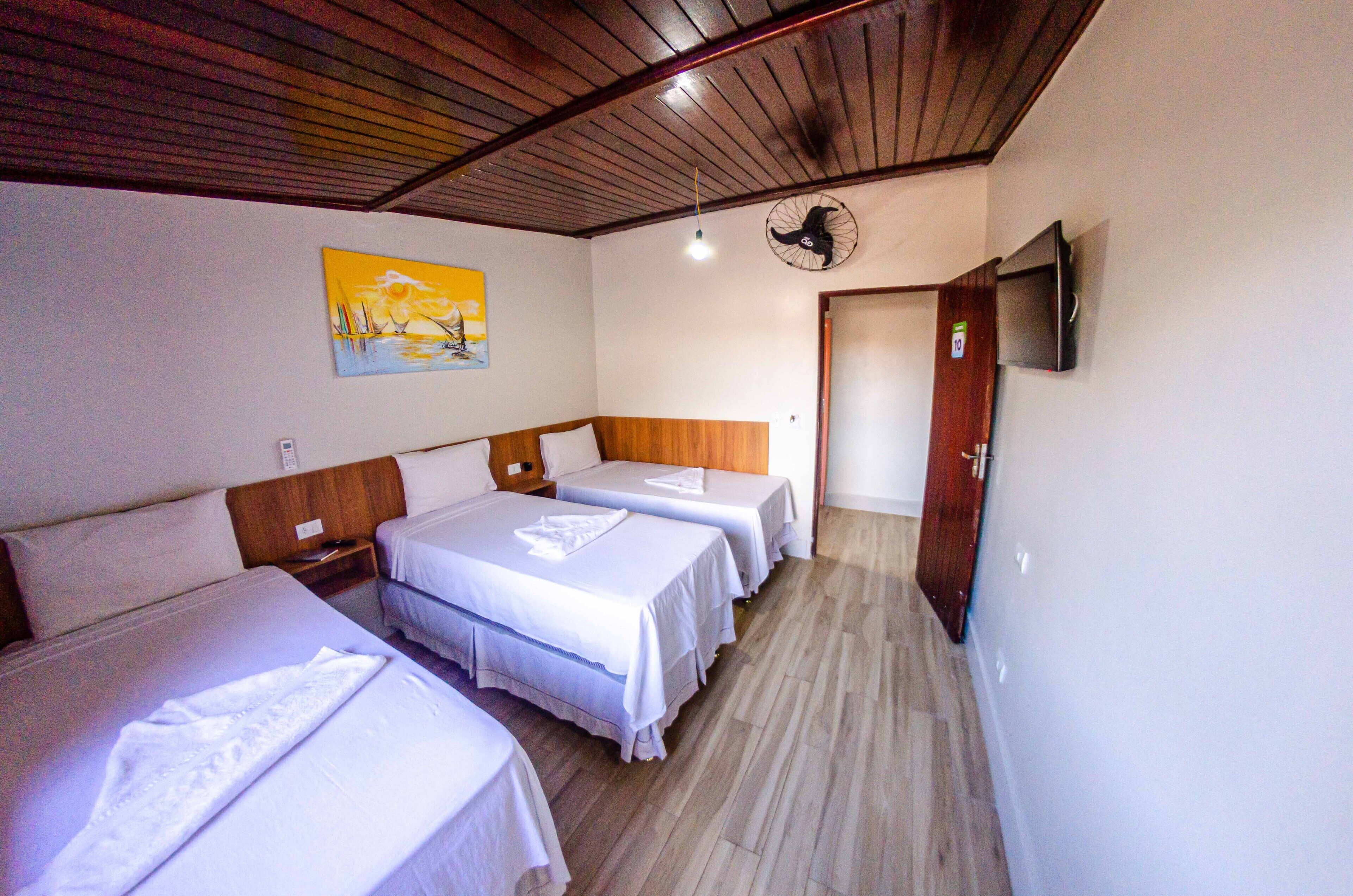A hotel room with three single beds, wooden headboards, a ceiling fan, a painting on the wall, and a TV mounted on the wall.