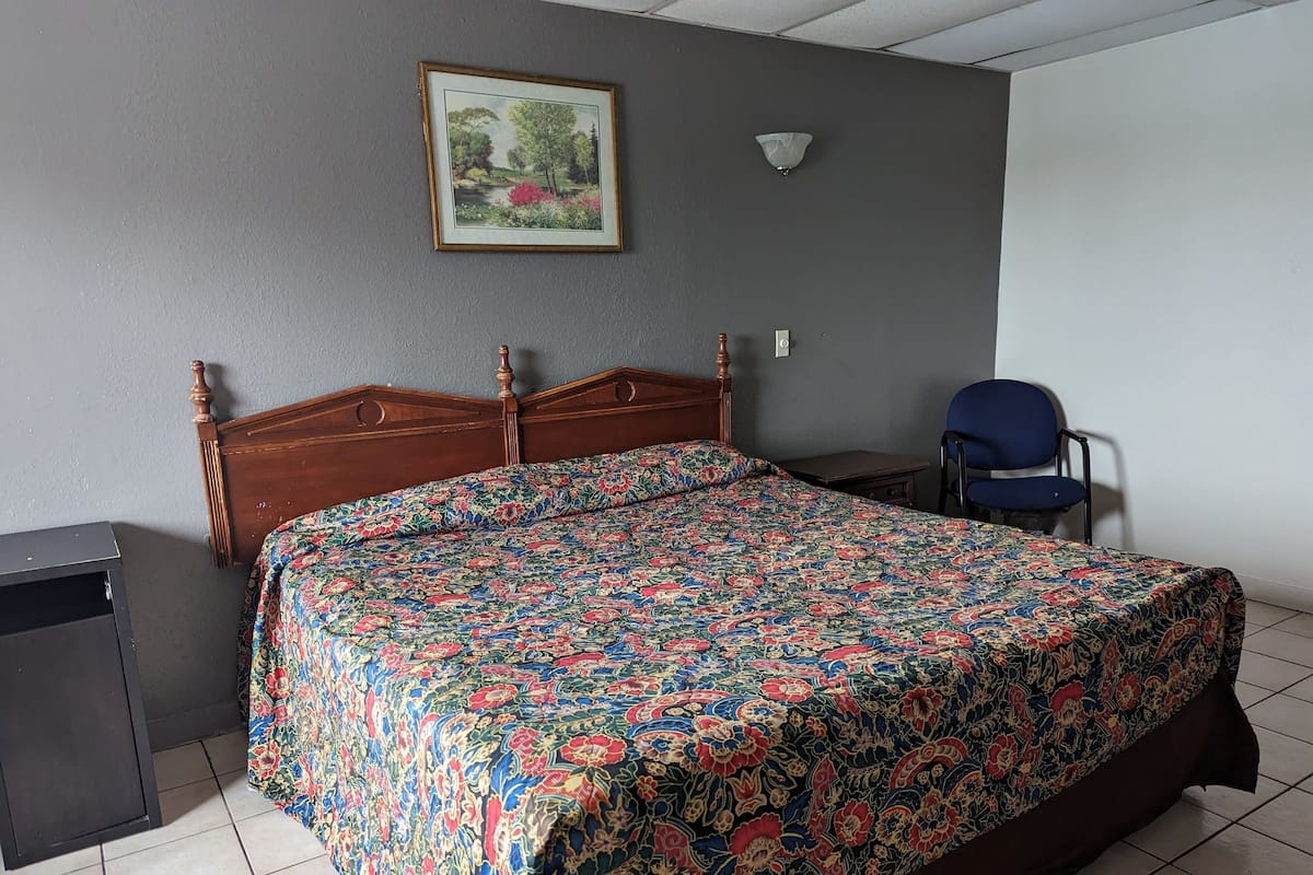 Una camera da letto con un letto di legno, una poltrona blu, un tavolino, un quadro appeso al muro e un pavimento piastrellato.