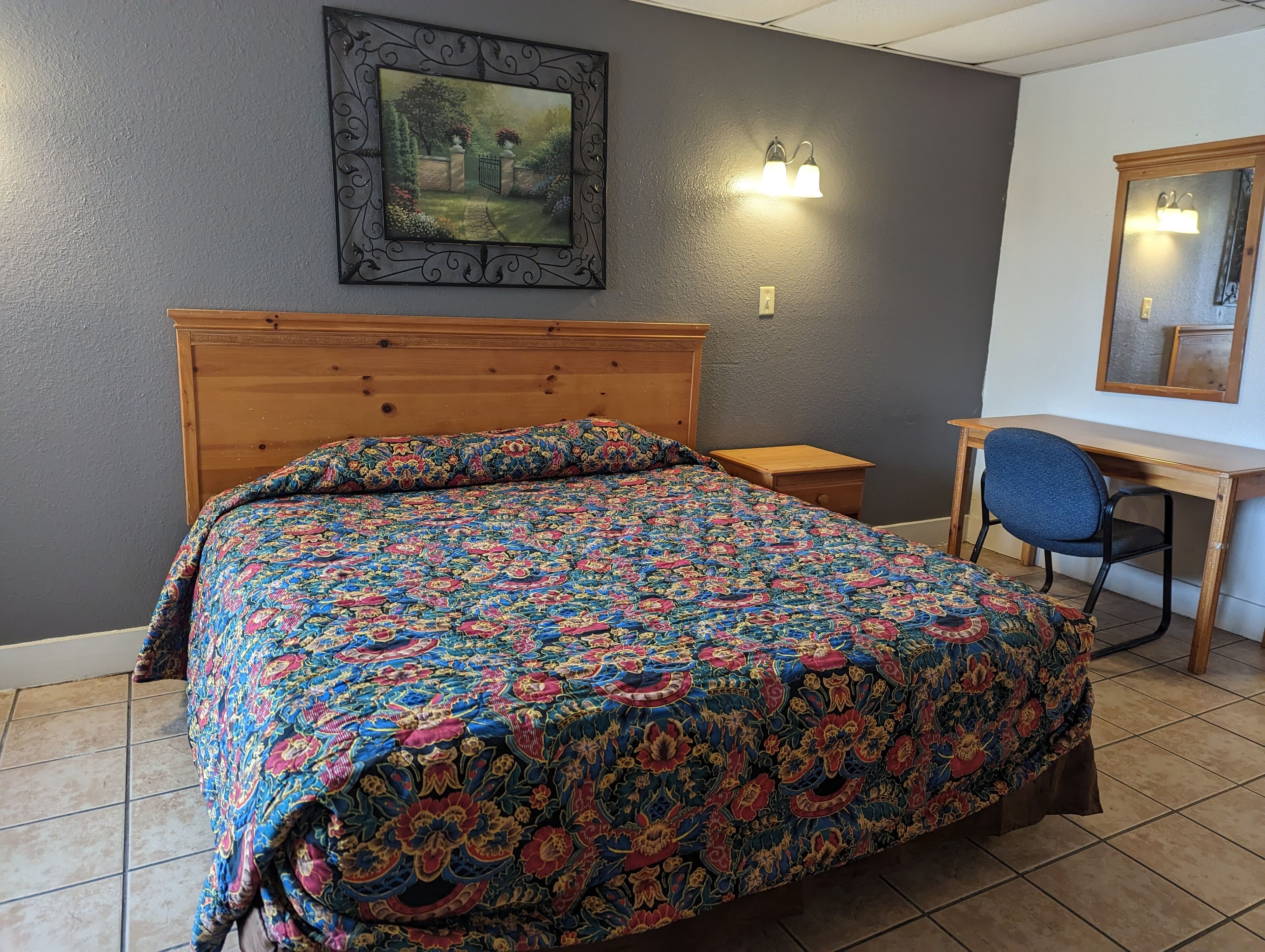 A bedroom with a wooden headboard, a colorful floral bedspread, a desk with a chair, and a wall-mounted painting.