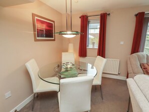 A dining area with a glass table surrounded by white chairs, a hanging light fixture, and a window with red curtains.