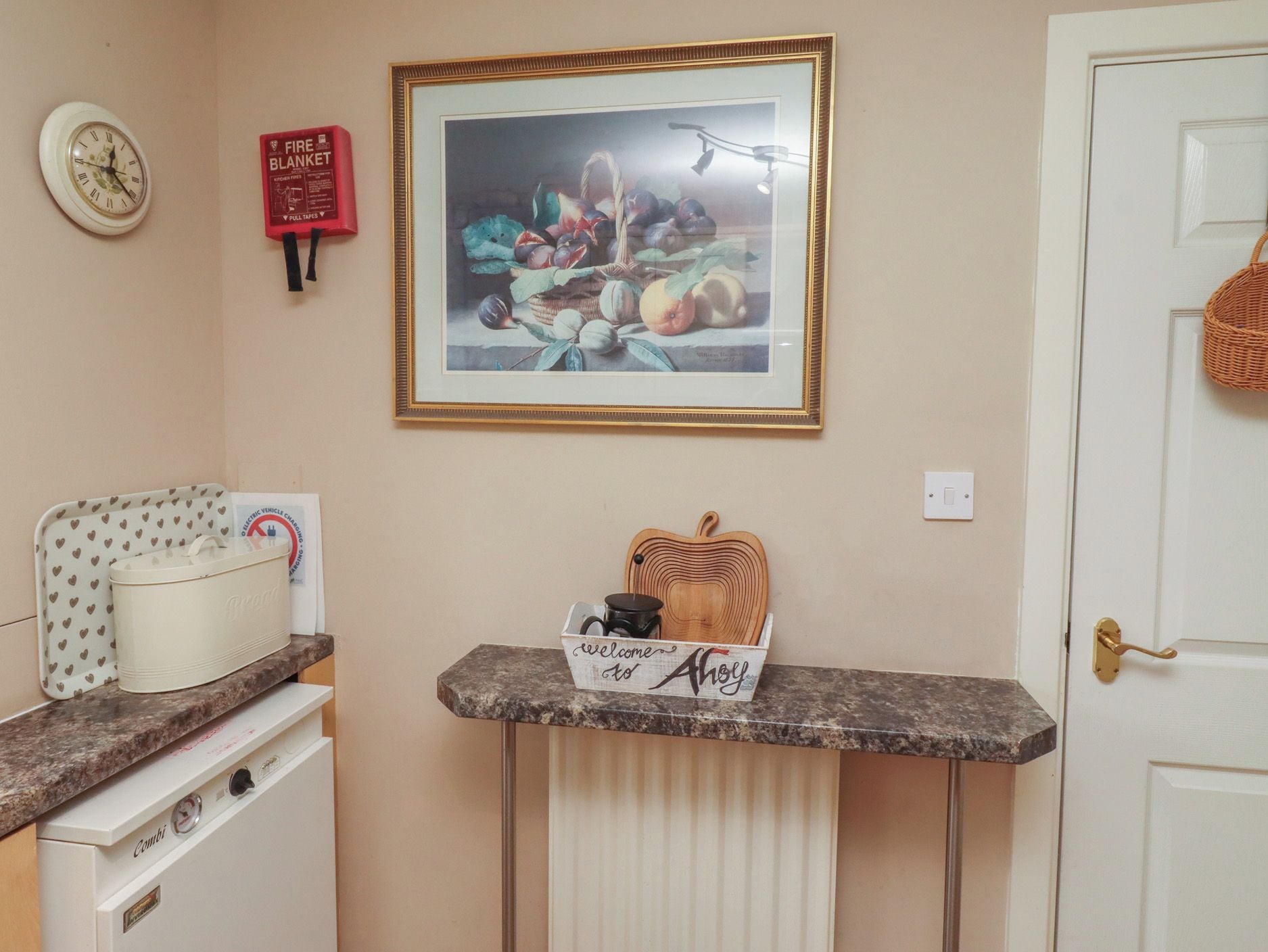 A kitchen area with a stove, a framed fruit still life, a fire blanket, and a welcome sign.