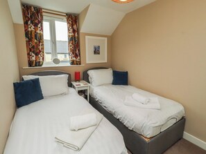 A bedroom with two beds, a bedside table, a red lamp, and a window with curtains. - Ahoy (Seahouses)