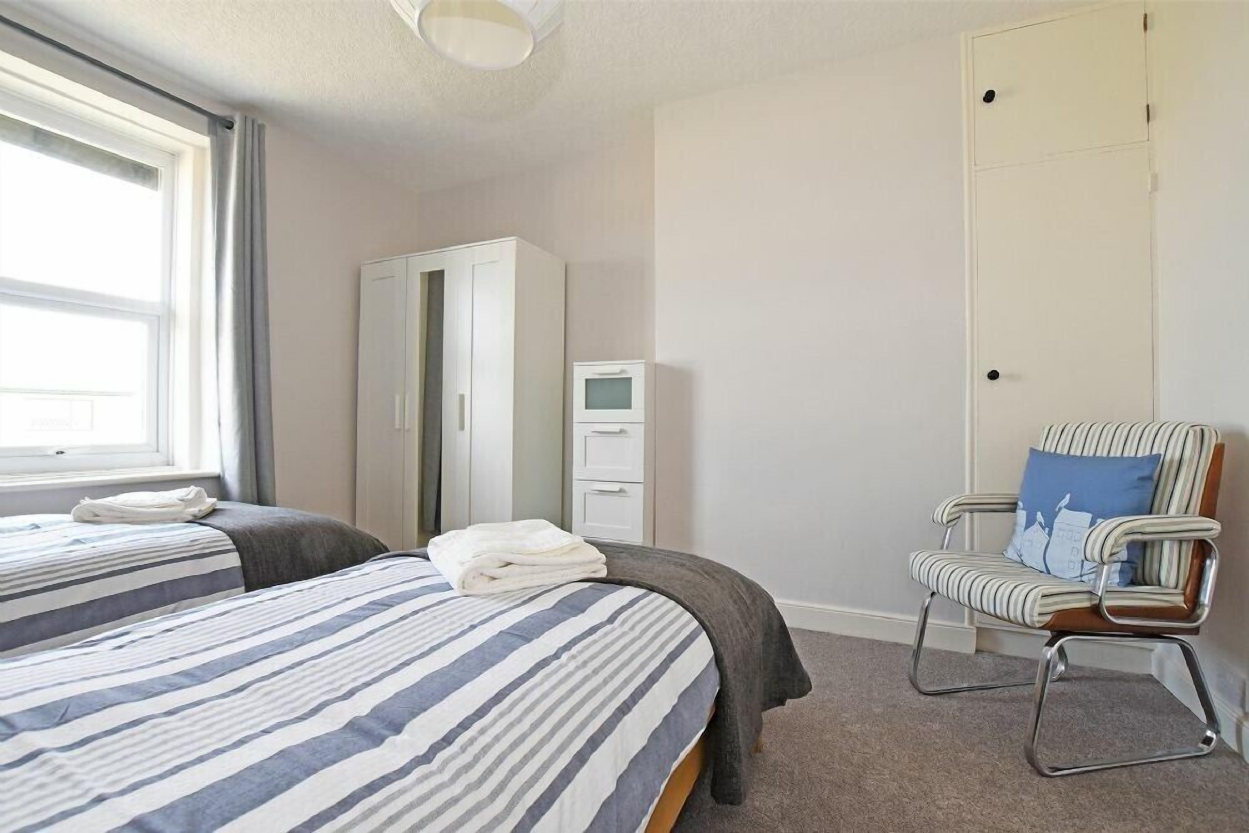 A hotel room with two beds, a wardrobe, a chair, and a small desk with a monitor.