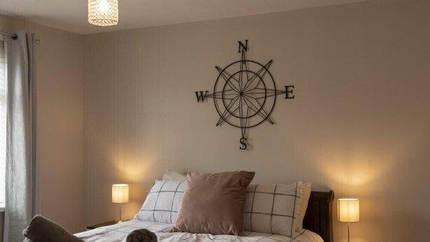 A neatly made bed with a compass wall decoration, two bedside lamps, and a window with curtains. - Southfield Lodge (Seahouses)
