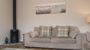 A living room with a beige sofa, a black wood-burning stove, and a painting of a coastal scene. - Southfield Lodge (Seahouses)