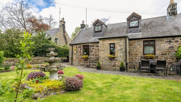 A stone house with a well-maintained garden, including a stone fountain and various plants. - Aryarch (Alnwick)
