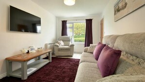 A living room with a flat-screen TV, a wooden coffee table, a grey sofa with purple cushions, a patterned armchair, and a window with curtains. - The Chevy (Seahouses)