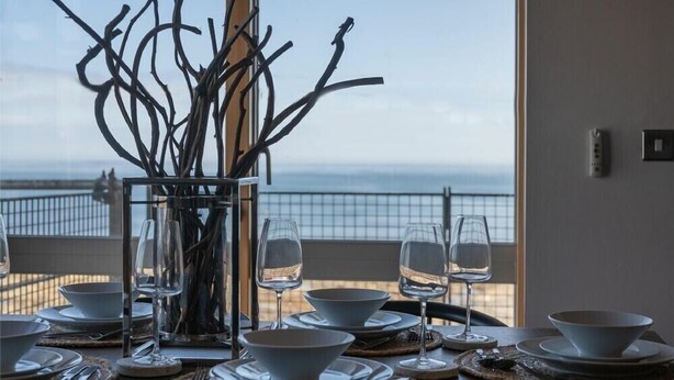 Tavola imbandita con piatti bianchi, ciotole e bicchieri da vino, con vista sul mare attraverso una finestra.