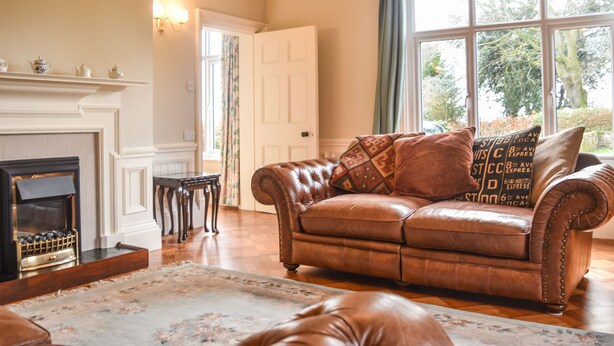 A living room with a leather sofa, a fireplace, and a large window with a view of trees. - Garden House (Alnwick)