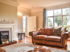 A living room with a leather sofa, a fireplace, and a large window with a view of trees. - Garden House (Alnwick)