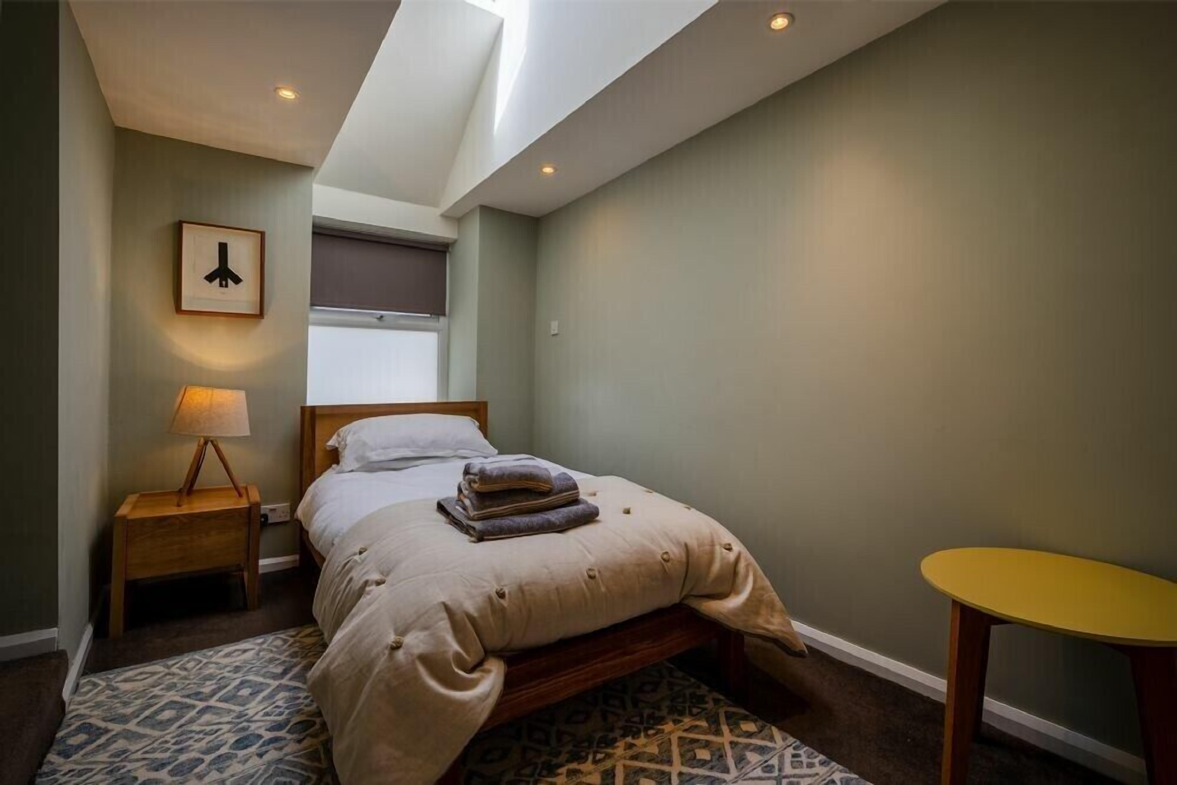 Un dormitorio con una cama, una mesita de noche con lámpara, una pequeña mesa redonda y una ventana con persianas.