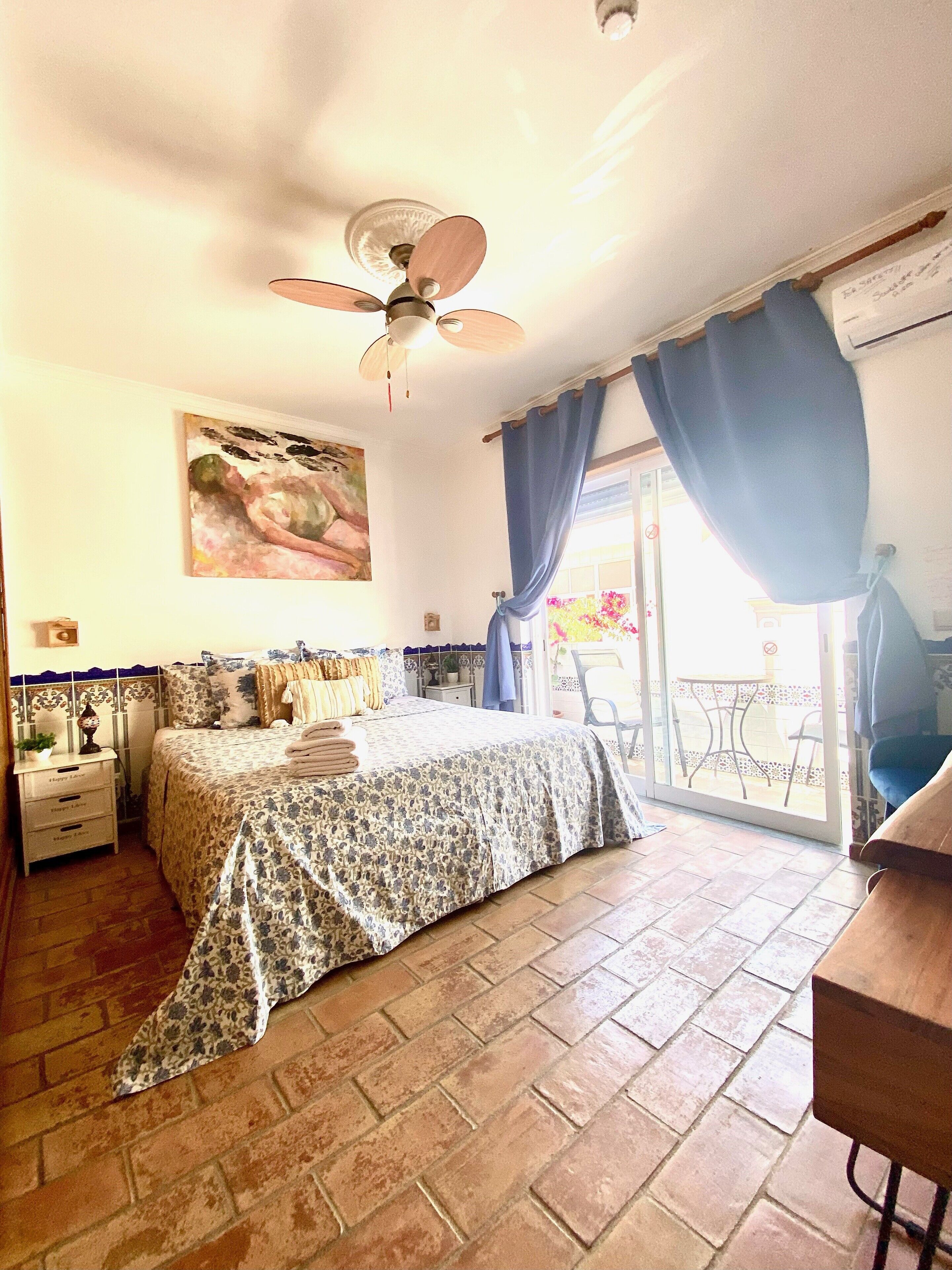 A bedroom with a bed, a ceiling fan, a painting, a nightstand, a window with curtains, and a view of the outside.