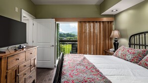 A bedroom with a bed, a TV, a dresser, a lamp, and a view of greenery through the window.