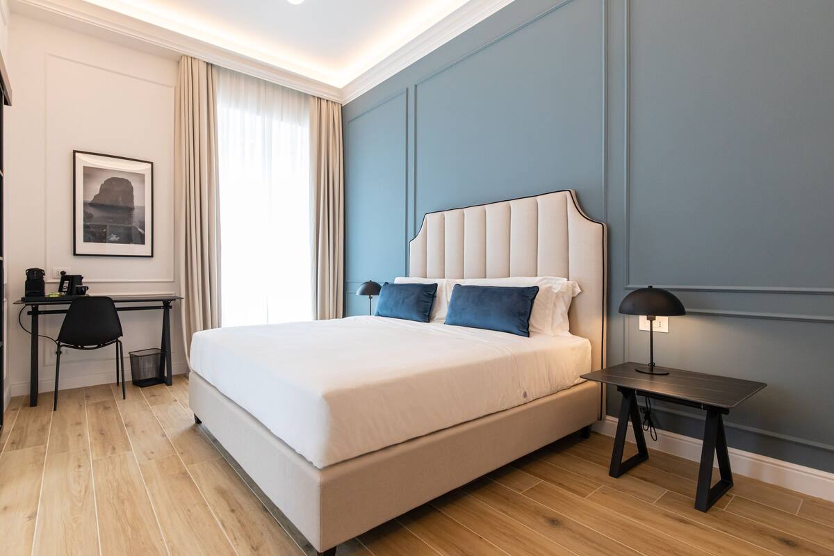 A modern hotel room with a large bed, a desk with a chair, a small table with a lamp, and a framed picture on the wall.
