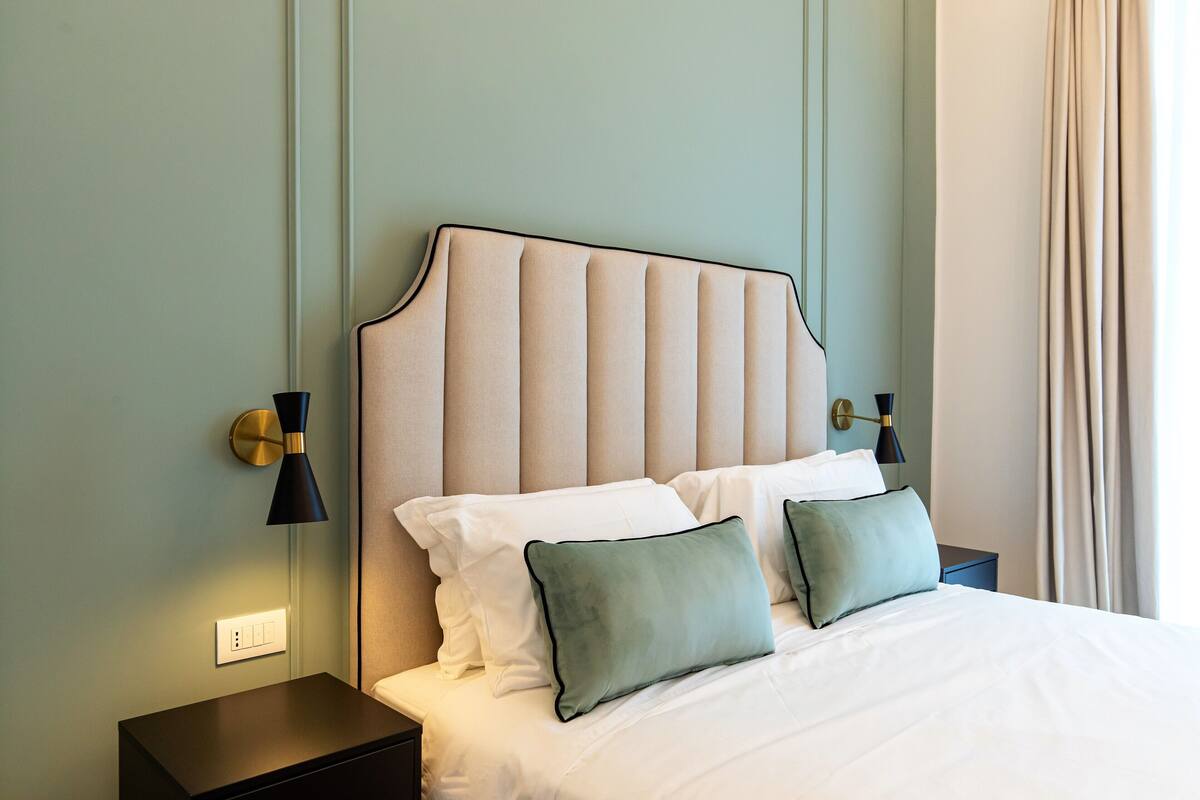 A neatly made bed with white linens and two green pillows, a bedside table with a lamp, and a wall-mounted light fixture.