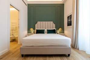 Uma cama bem arrumada com cabeceira, duas luminárias de apoio e vista para um banheiro com pia de mármore e banheira.
