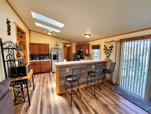 Una cocina con armarios de madera, una isla central con taburetes de bar, un refrigerador y un ventanal con persianas.