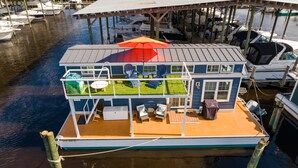 Een woonboot met een dek, buitenmeubels en een overdekt terras met stoelen en een parasol.