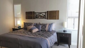 Quarto com uma cama grande, duas mesas de cabeceira, um prateleira na parede e uma janela com persianas.