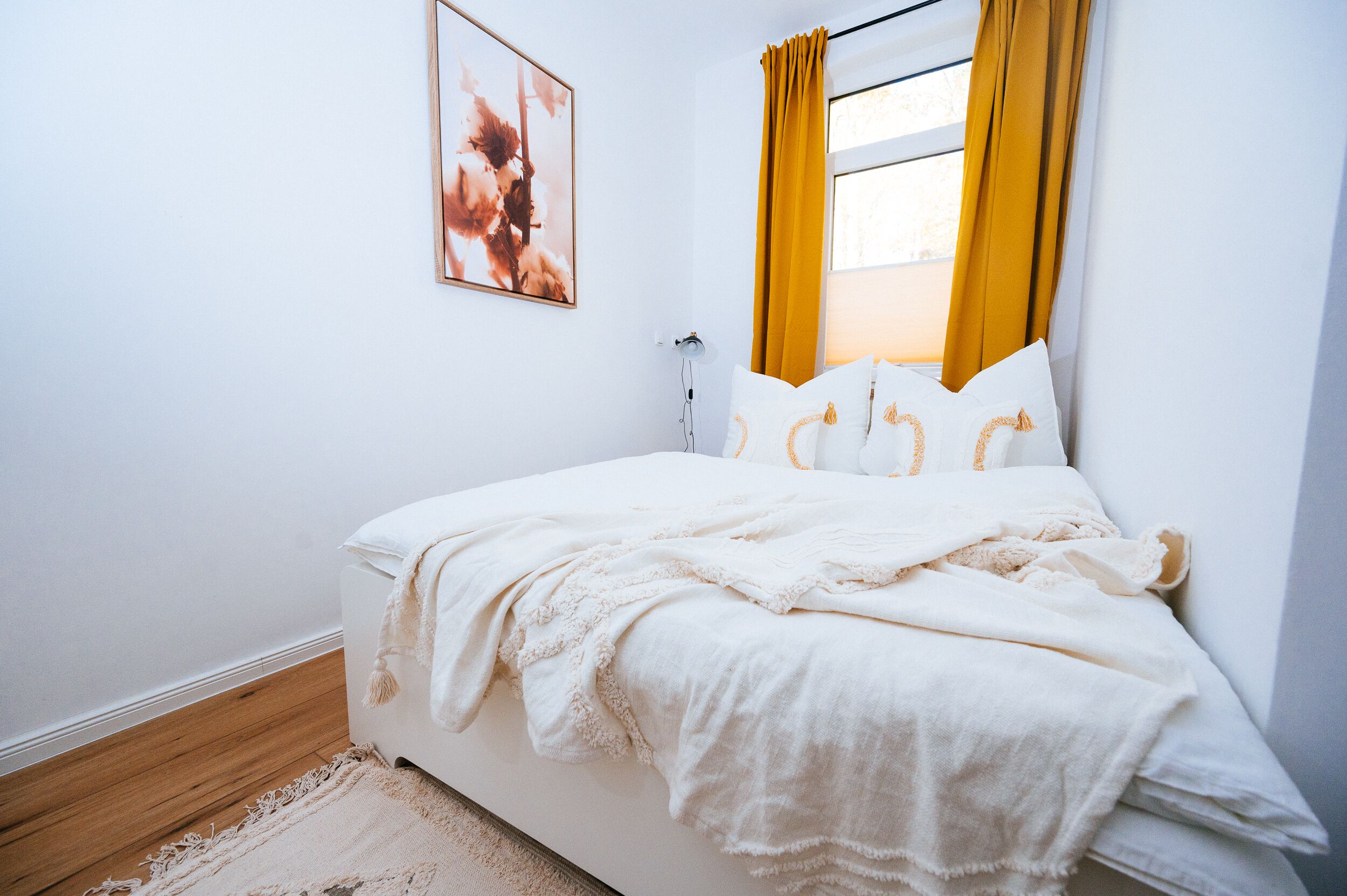 Una cama bien hecha con sábanas blancas y un plaid color beige, una mesita de noche con una lámpara, y una ventana con cortinas amarillas.