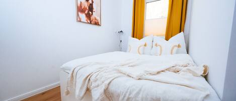 Una cama bien hecha con sábanas blancas y un plaid color beige, una mesita de noche con una lámpara, y una ventana con cortinas amarillas.