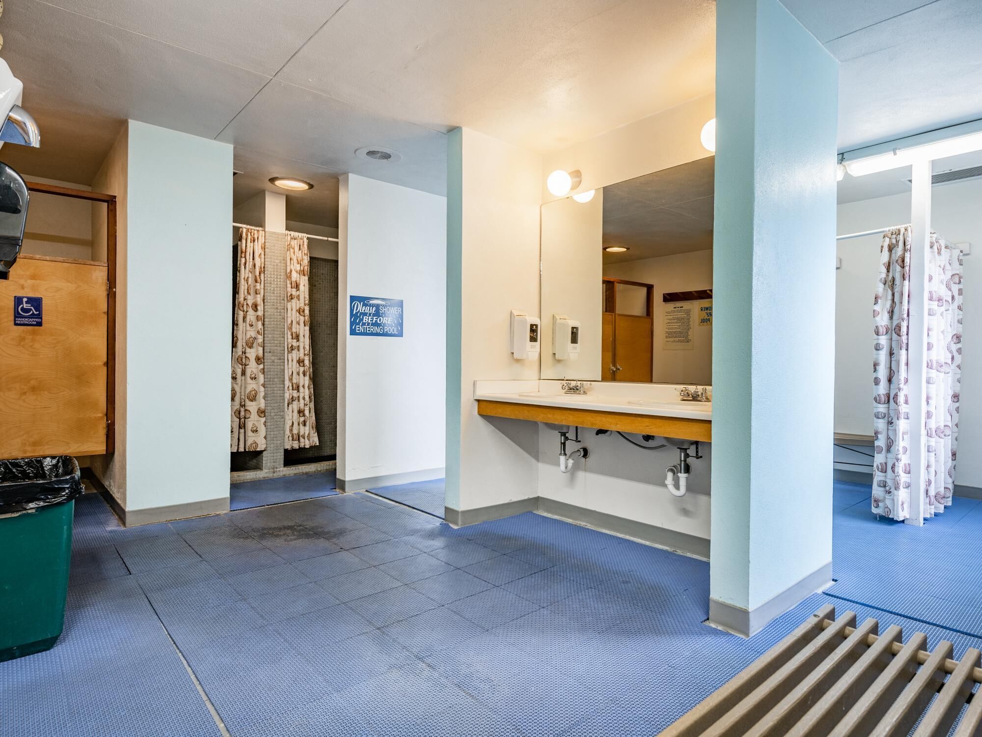 A public restroom with a blue floor, a hand dryer, a mirror, and a sign instructing to shower before entering the pool.