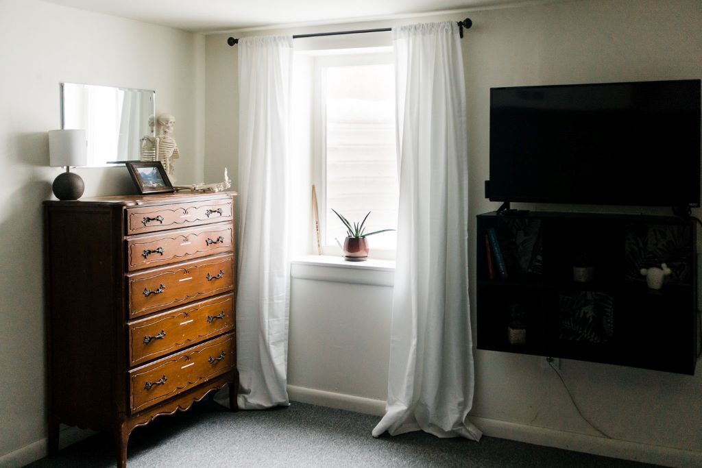 Quarto com cômoda de madeira, televisão fixada na parede e janela com cortinas brancas.