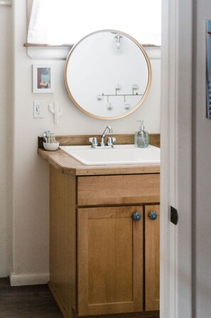 A bathroom with a wooden vanity, a round mirror, a white sink, and a faucet. - Downtown Bozeman Bungalow - 2 blocks from Main St. (Bozeman)