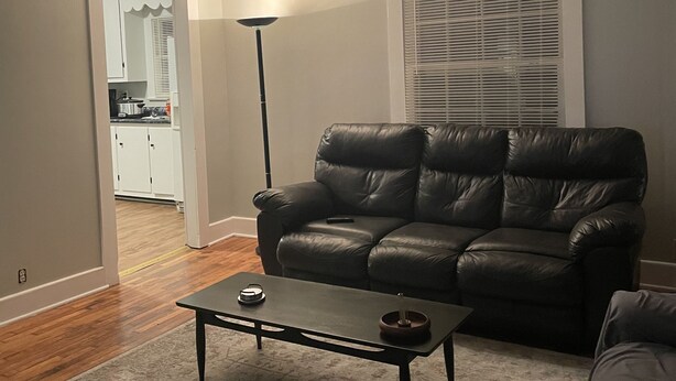 A living room with a black leather sofa, a coffee table, and a floor lamp. There is a kitchen visible through an open doorway.
