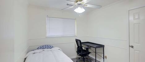Une chambre à coucher comprenant un lit, un bureau, une chaise, un ventilateur de plafond et une fenêtre avec des stores.
