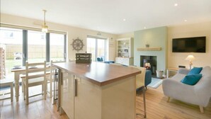 A modern kitchen with a central island, a dining area with a wooden table and chairs, a living room with a sofa and a fireplace, and a TV mounted on the wall. - Beach Walk (Chathill)