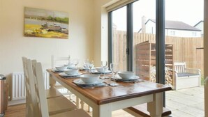 A dining area with a wooden table set for six, white chairs, and a large window offering a view of a backyard. - Beach Walk (Chathill)