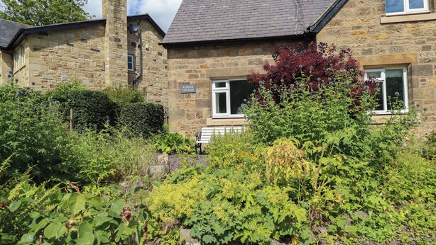 A stone cottage with a garden in front, a bench, and a sign reading "AINSIDE COTTAGE". - Alnside Cottage (Alnwick)