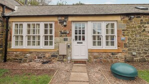 A stone cottage with a white door, two windows with shutters, and a small garden area with a pathway leading to the entrance. - Coral Cottage (Bamburgh)