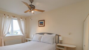 A bedroom with a bed, a ceiling fan, a window with curtains, a bedside table with a lamp, and a wall-mounted picture.